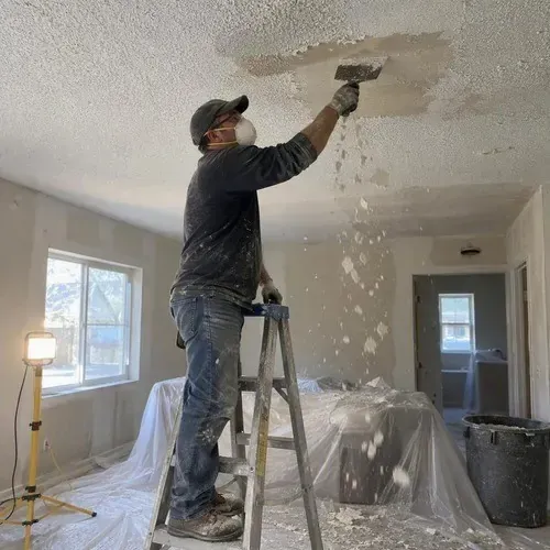 Masked drywall worker scraping popcorn stipple ceiling texture inside plastic containment in Kingston